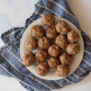 A white plate with several round, brown chocolate peanut butter protein balls topped with oats. The plate is placed on a crumpled, blue and white striped cloth on a marble surface.