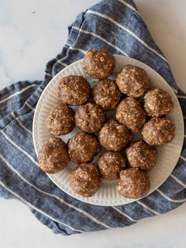 A white plate with several round, brown chocolate peanut butter protein balls topped with oats. The plate is placed on a crumpled, blue and white striped cloth on a marble surface.