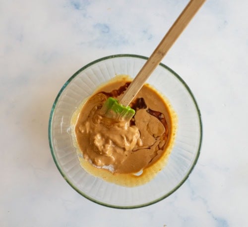 A glass bowl containing a mixture of creamy peanut butter, soy sauce, and a green spatula resting inside. The ingredients are partially blended, creating a marbled effect on a light marble surface.