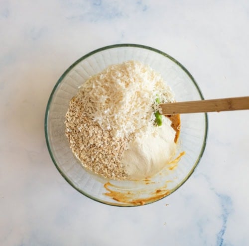 A glass bowl containing oats, shredded coconut, flour, and a brown liquid is being mixed with a wooden spoon on a light surface.