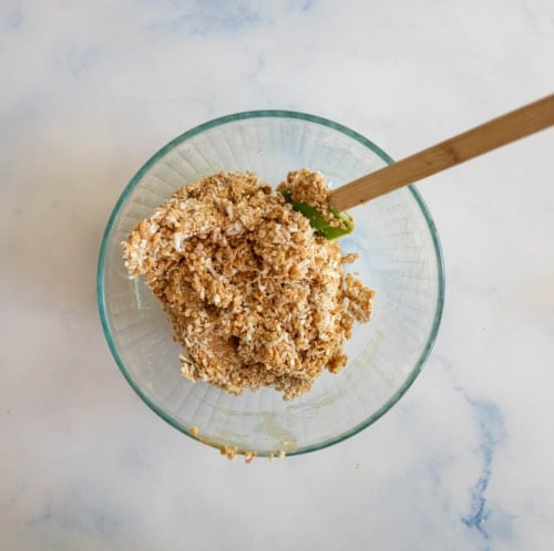 A glass bowl on a light surface contains a mixture of oats and shredded coconut. A wooden spatula with a green silicone end is mixing the ingredients.
