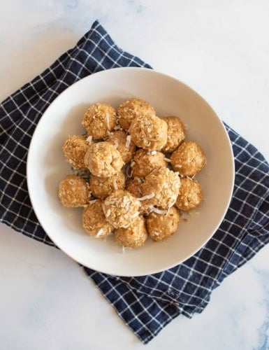 A white bowl filled with energy balls topped with shredded coconut is placed on a dark blue checkered cloth. The background is a light marble surface.