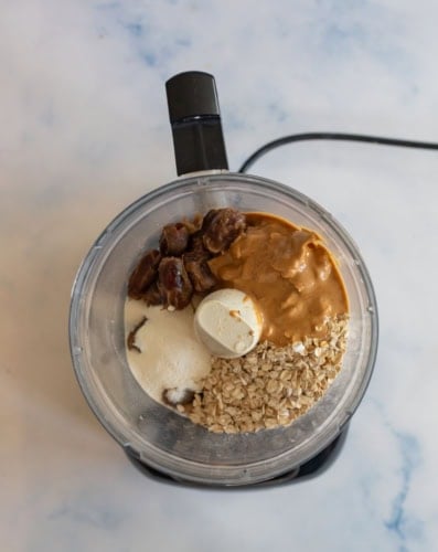 Overhead view of a food processor containing rolled oats, peanut butter, dates, and protein powder on a light blue marbled surface.