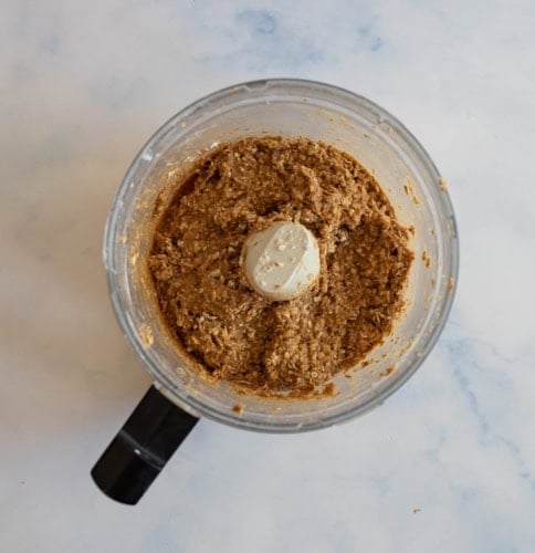Top view of a food processor containing a brown, coarse mixture. The mixture appears to be a blend of ingredients, likely a batter or dough, with visible textures of nuts or grains. The processor lid and handle are visible.