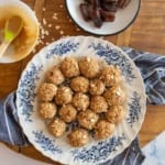 A plate with medjool date protein bites arranged in a circle, placed on a floral-patterned dish. Nearby, a bowl of dates and a bowl with a spoon and peanut butter are on a wooden surface with a striped cloth.