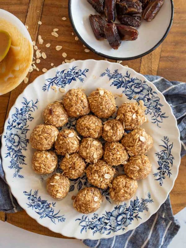A plate with medjool date protein bites arranged in a circle, placed on a floral-patterned dish. Nearby, a bowl of dates and a bowl with a spoon and peanut butter are on a wooden surface with a striped cloth.