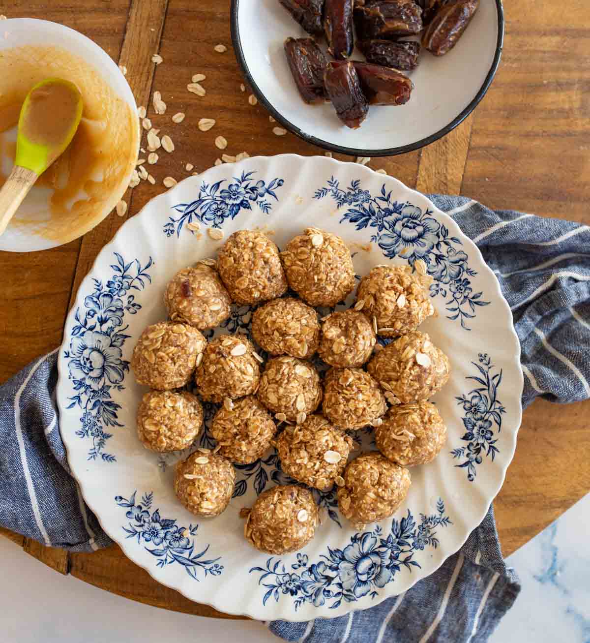 A plate with medjool date protein bites arranged in a circle, placed on a floral-patterned dish. Nearby, a bowl of dates and a bowl with a spoon and peanut butter are on a wooden surface with a striped cloth.