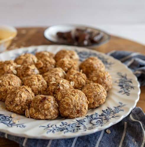 A decorative plate holds several round oat and date balls. The plate has blue floral patterns and is placed on a wooden surface. A striped cloth and a small dish with dates are in the background.