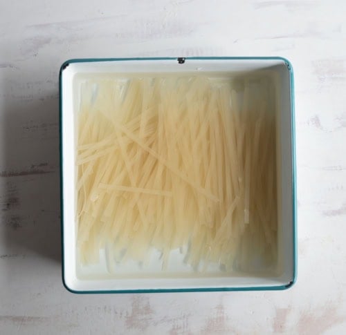 A square, white enamel tray filled with uncooked, translucent flat rice noodles soaking in water, on a light-colored surface.
