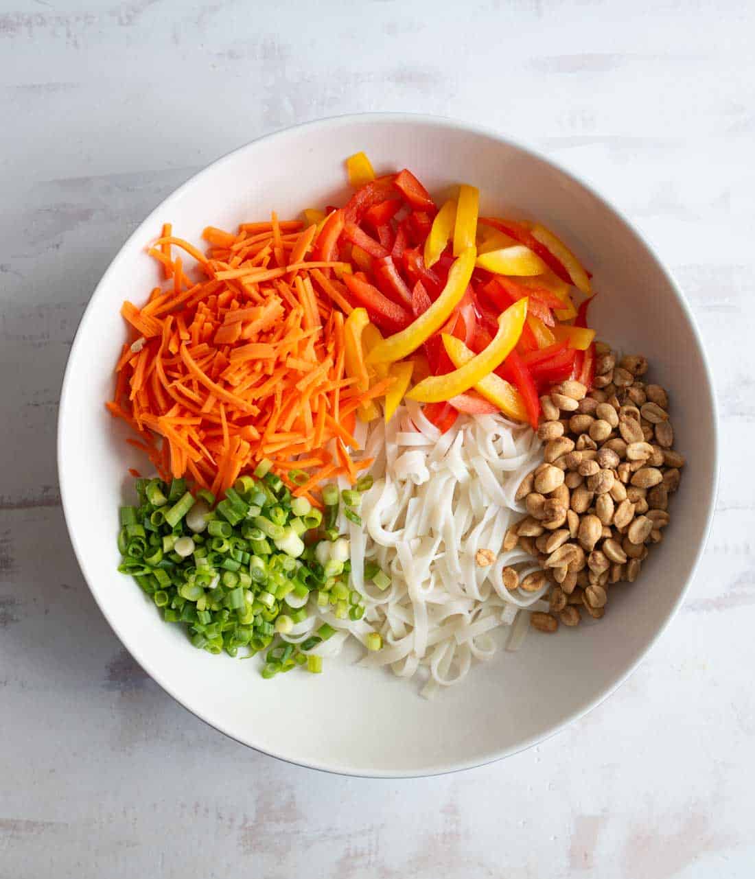 A white bowl filled with shredded carrots, sliced red and yellow bell pepper, chopped green onions, rice noodles, and peanuts, arranged in separate sections on a light surface.