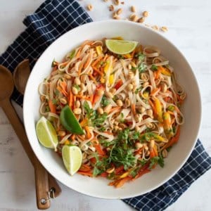 A white bowl of colorful Asian noodle salad with carrots, peppers, peanuts, cilantro, and lime wedges sits on a blue checkered napkin, with wooden utensils nearby. Some peanuts are scattered on the table.