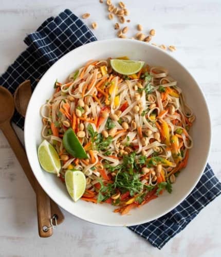 A white bowl of colorful Asian noodle salad with carrots, peppers, peanuts, cilantro, and lime wedges sits on a blue checkered napkin, with wooden utensils nearby. Some peanuts are scattered on the table.