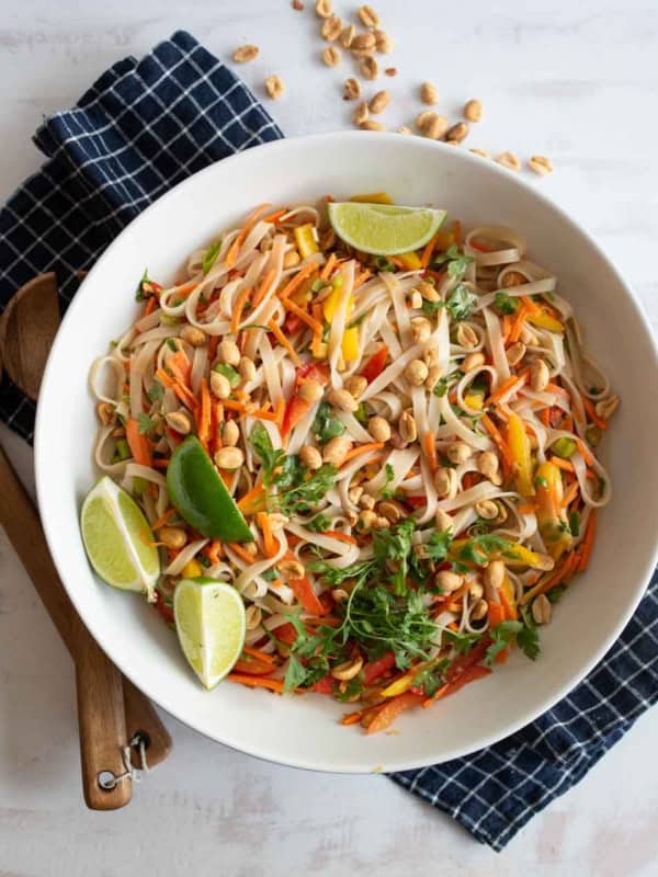 A white bowl of colorful Asian noodle salad with carrots, peppers, peanuts, cilantro, and lime wedges sits on a blue checkered napkin, with wooden utensils nearby. Some peanuts are scattered on the table.