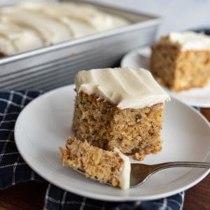A square slice of banana cake with cream cheese frosting sits on a white plate with a fork holding a bite, in front of a pan and another plate with more cake. A dark checkered cloth is underneath the plate.