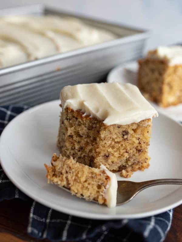 A square slice of banana cake with cream cheese frosting sits on a white plate with a fork holding a bite, in front of a pan and another plate with more cake. A dark checkered cloth is underneath the plate.