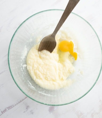 A glass bowl containing a mixture of wet and dry ingredients, with a cracked egg and a wooden spoon resting inside, on a white surface.