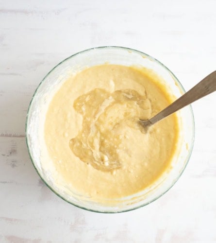 A glass bowl filled with yellow pancake batter sits on a light surface, with a metal spoon resting inside the bowl.