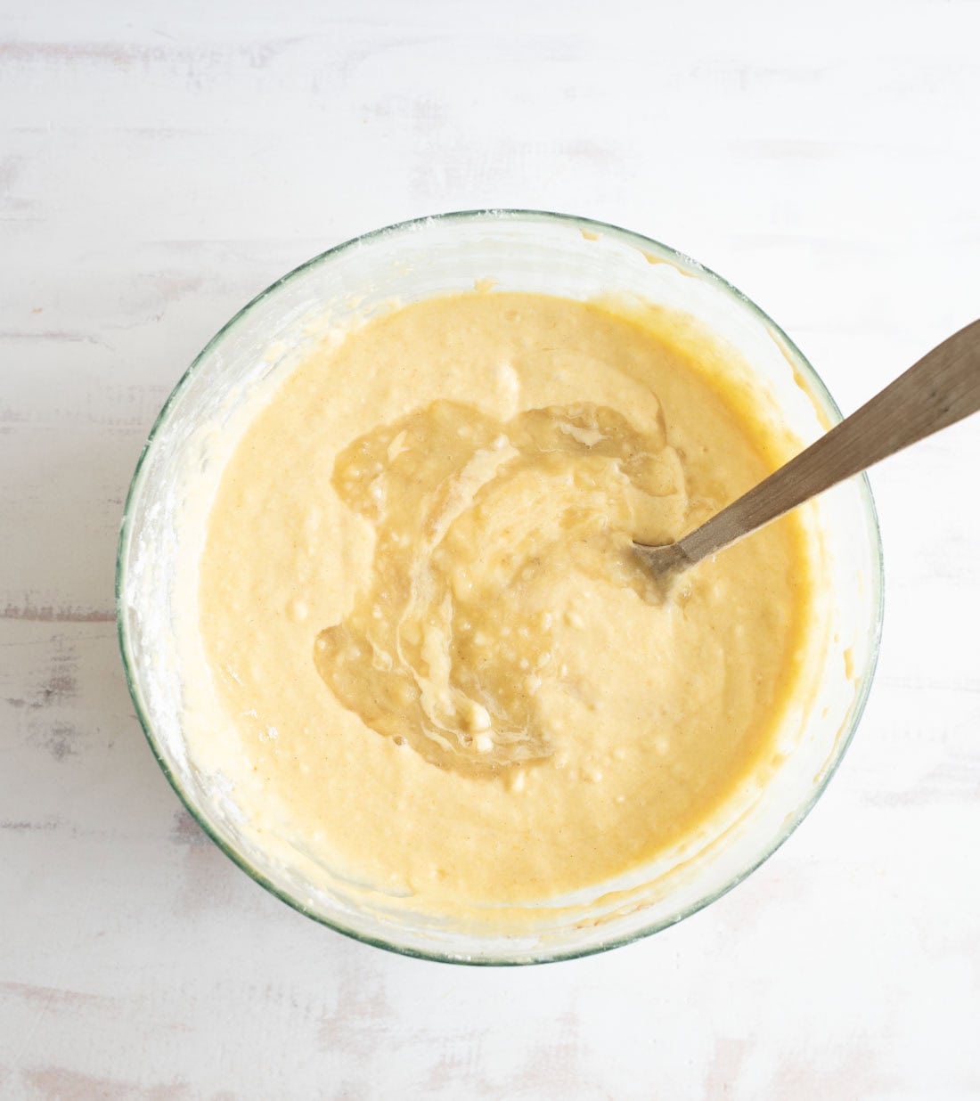 A glass bowl filled with yellow pancake batter sits on a light surface, with a metal spoon resting inside the bowl.