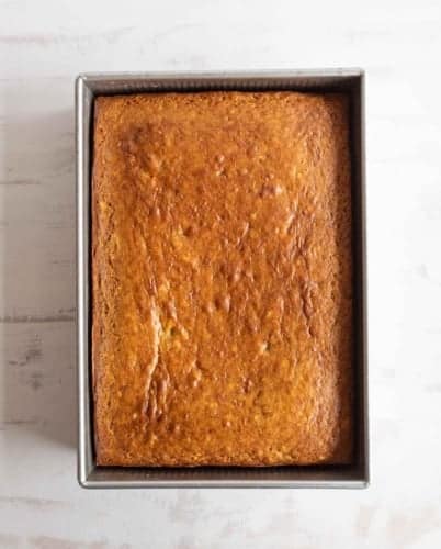 A golden-brown, rectangular loaf of baked banana bread sits in a metal baking pan on a light-colored surface.