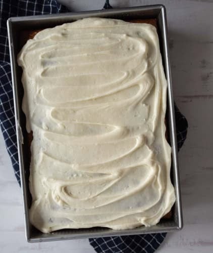A rectangular cake in a metal pan is topped with a thick layer of swirled white frosting. The pan sits on a dark, checkered cloth against a light surface.