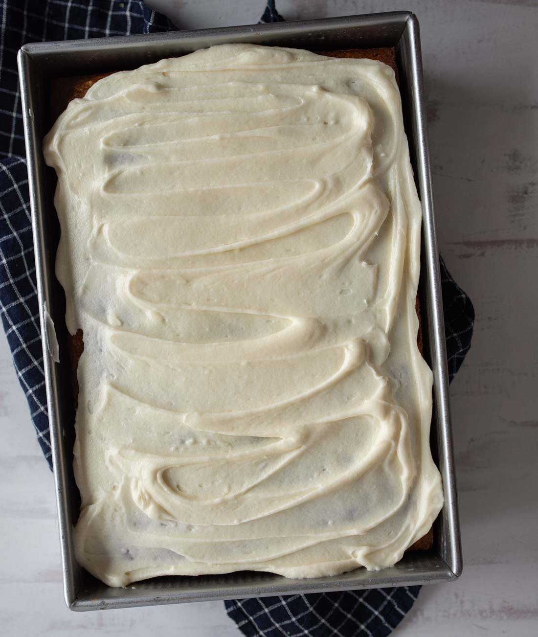 A rectangular cake in a metal pan is topped with a thick layer of swirled white frosting. The pan sits on a dark, checkered cloth against a light surface.