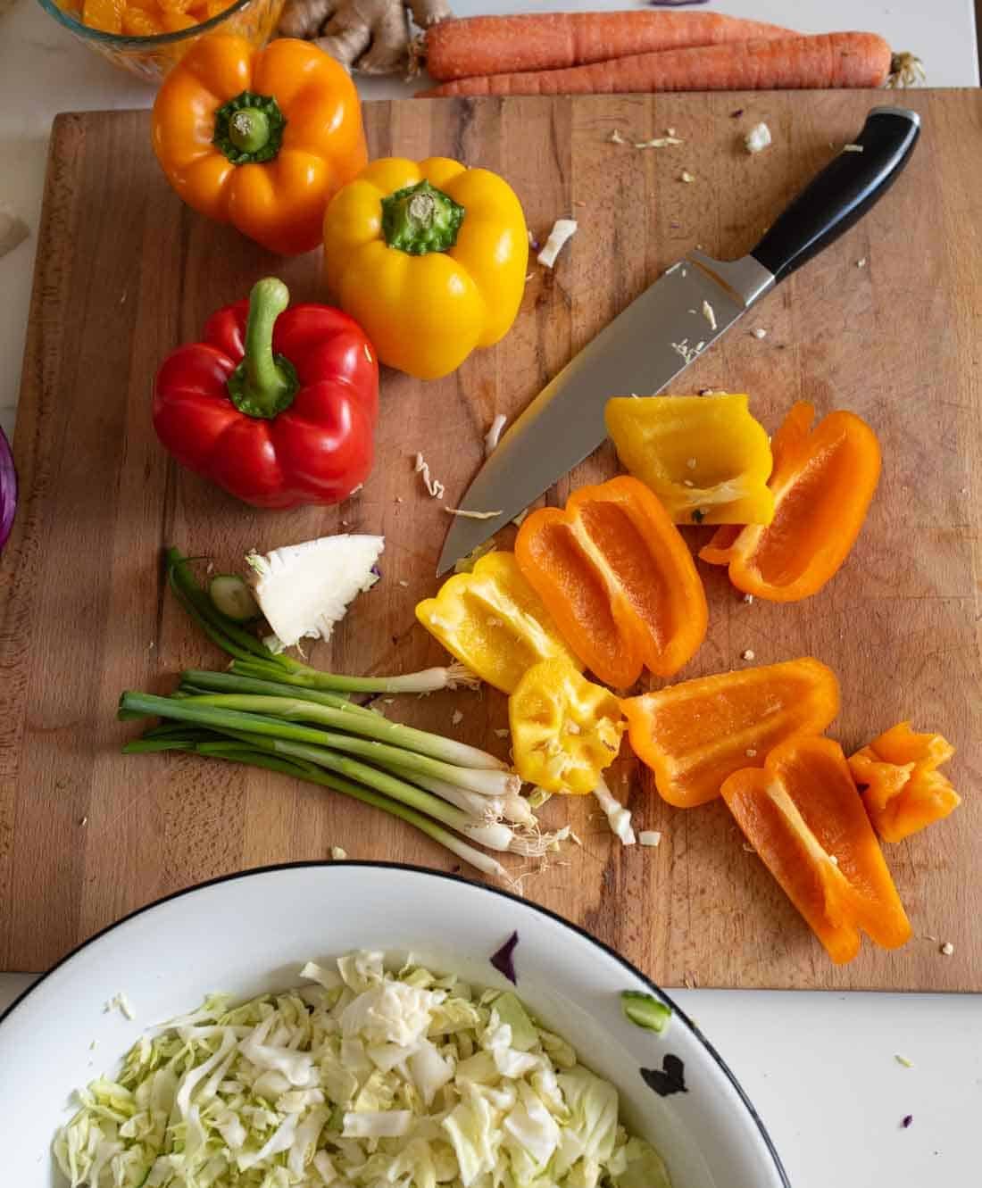 Chopped orange and yellow bell peppers, green onions, a knife, and whole bell peppers are on a wooden cutting board, with carrots and ginger nearby. A bowl of shredded cabbage sits at the bottom.