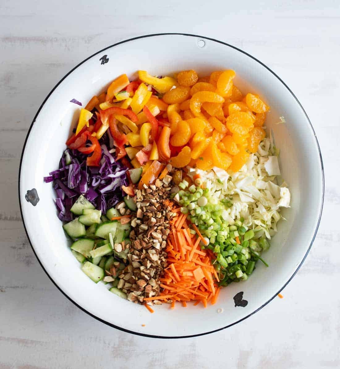 A white bowl filled with colorful ingredients for a salad, including sliced red and yellow bell peppers, mandarin orange segments, chopped green onions, shredded carrots, cucumber, purple cabbage, white cabbage, and chopped nuts.