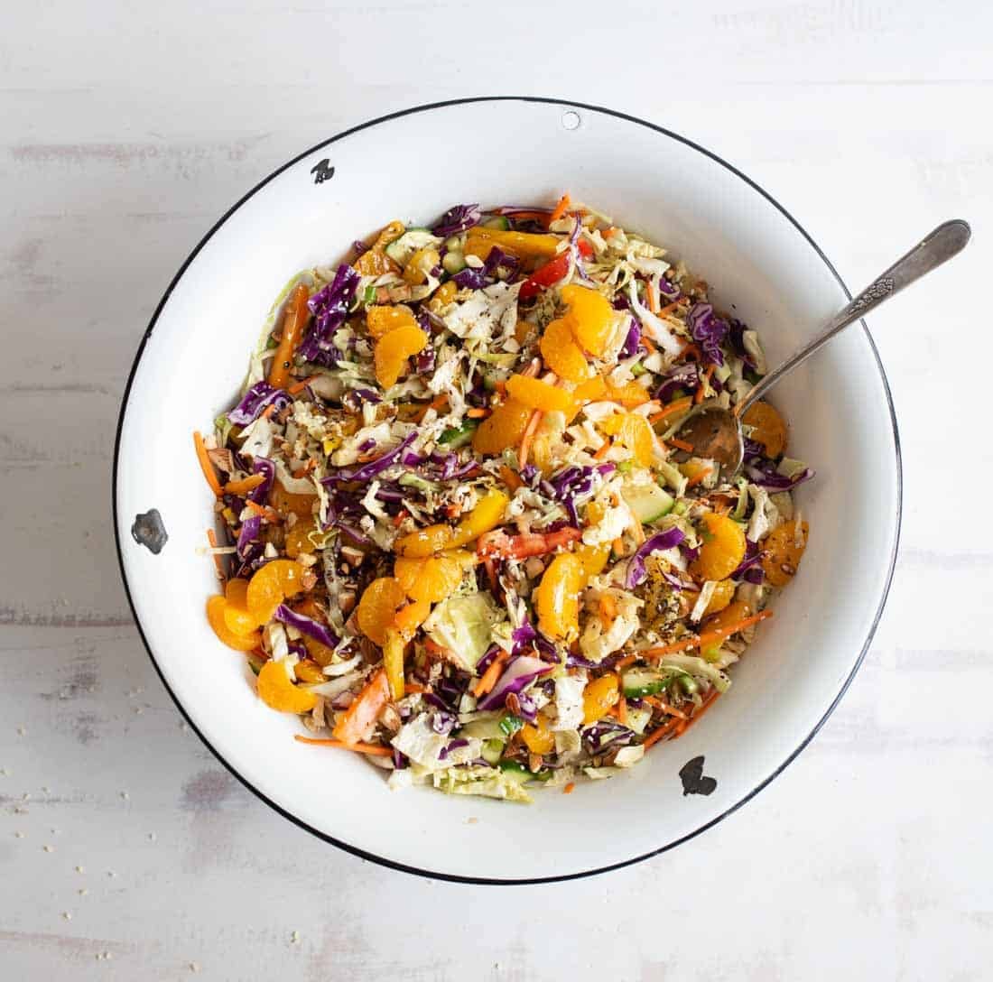 A large white bowl filled with colorful Asian-inspired salad featuring shredded cabbage, carrots, purple cabbage, sliced green onions, mandarin orange segments, and a sprinkling of sesame seeds, with a spoon in the bowl.