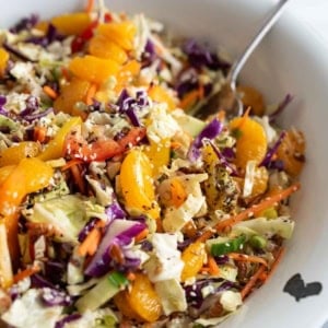 Colorful cabbage salad with shredded carrots, red cabbage, green cabbage, bell peppers, orange segments, and seeds, tossed together in a white bowl with a spoon.
