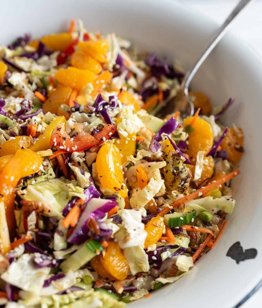 Colorful cabbage salad with shredded carrots, red cabbage, green cabbage, bell peppers, orange segments, and seeds, tossed together in a white bowl with a spoon.