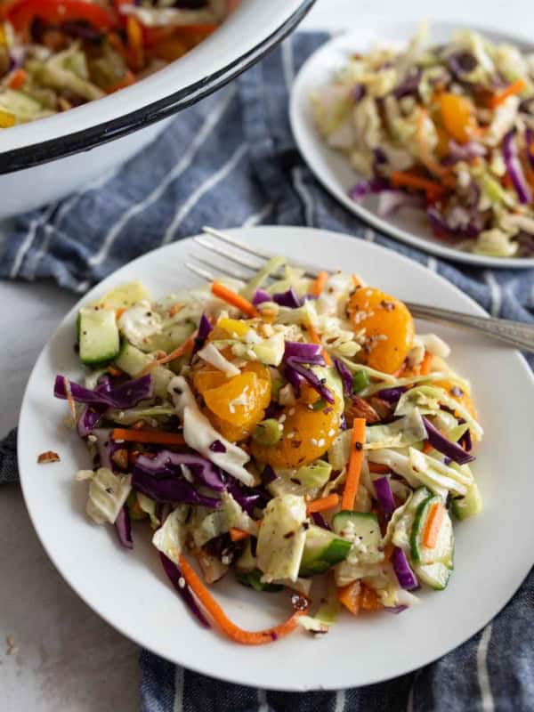 A plate of colorful Asian cabbage salad with shredded carrots, cucumbers, cherry tomatoes, and sesame seeds, served with a fork on a blue-striped cloth. Another plate and bowl of salad are in the background.