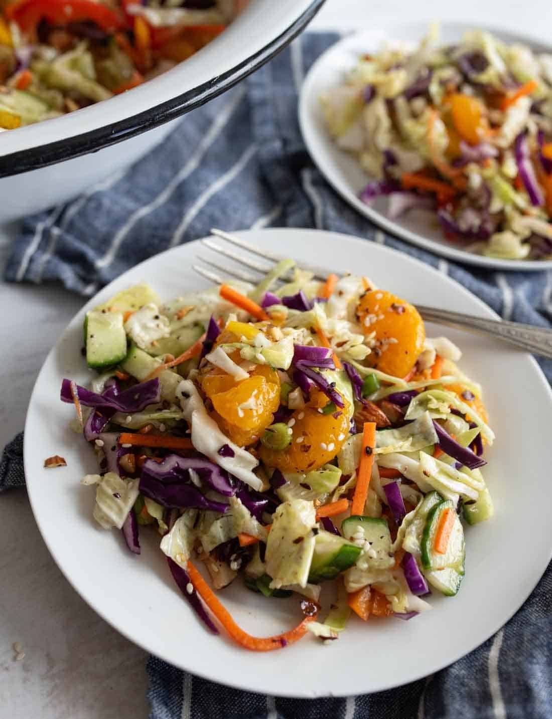 A plate of colorful Asian cabbage salad with shredded carrots, cucumbers, cherry tomatoes, and sesame seeds, served with a fork on a blue-striped cloth. Another plate and bowl of salad are in the background.