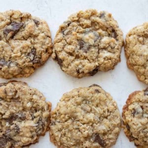 Six oatmeal chocolate chip cookies are arranged on a light-colored surface. The cookies appear chewy and have visible oats and chunks of chocolate throughout.