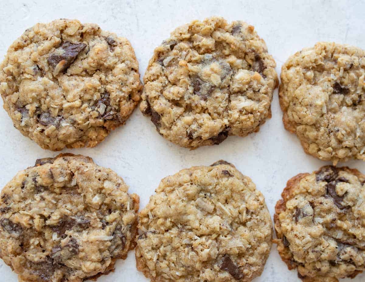 Six oatmeal chocolate chip cookies are arranged on a light-colored surface. The cookies appear chewy and have visible oats and chunks of chocolate throughout.