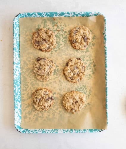 A blue speckled baking tray lined with parchment paper holds six oatmeal chocolate chip cookies arranged in two columns of three on a light-colored surface.