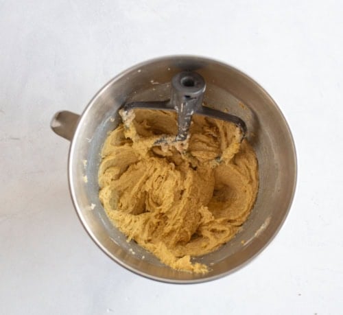A mixing bowl with a paddle attachment, filled with light brown cookie dough, sits on a white surface.