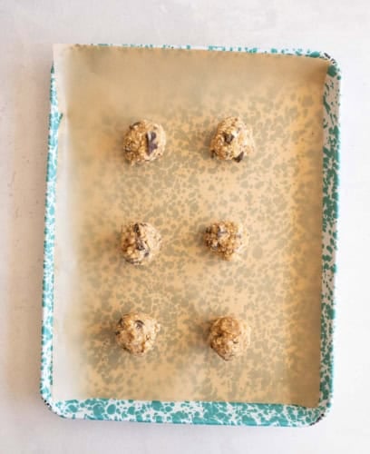 A teal-speckled baking sheet lined with parchment paper holds six evenly spaced balls of cookie dough, ready to be baked.