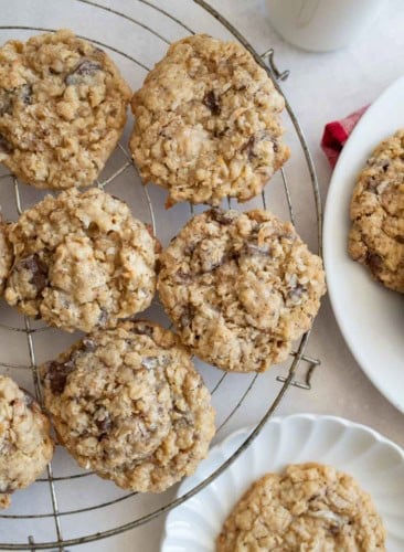 A cooling rack with several oatmeal coconut cookies, with more cookies on a white plate nearby. The cookies are golden brown and chunky, sitting on a light-colored surface.