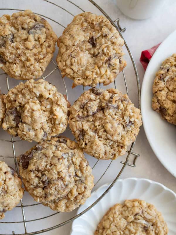 A cooling rack with several oatmeal coconut cookies, with more cookies on a white plate nearby. The cookies are golden brown and chunky, sitting on a light-colored surface.