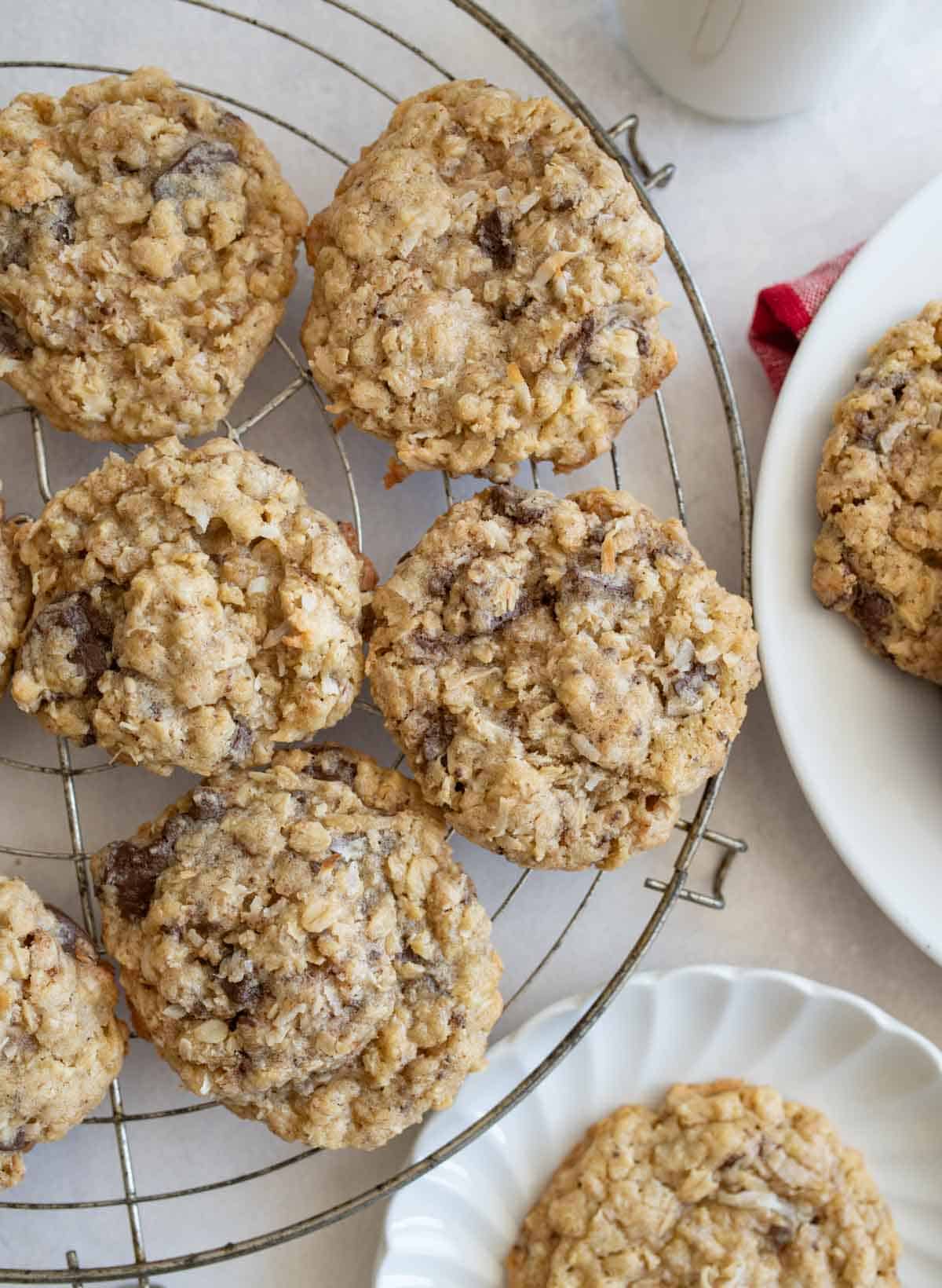 A cooling rack with several oatmeal coconut cookies, with more cookies on a white plate nearby. The cookies are golden brown and chunky, sitting on a light-colored surface.