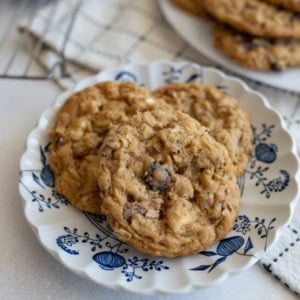 Three homemade oatmeal cookies with chocolate chips and nuts on a decorative white and blue floral plate, placed on a light surface with a checkered cloth and more cookies in the background.