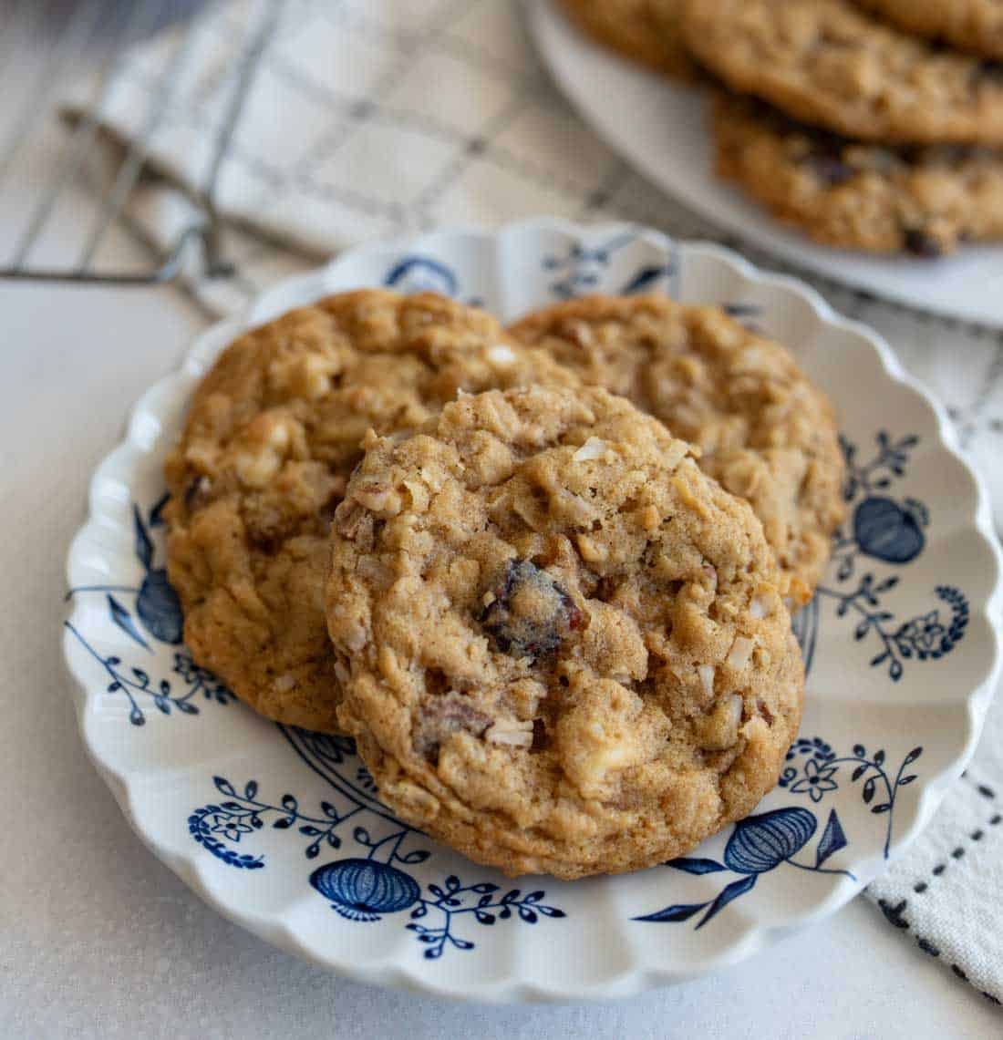 Three homemade oatmeal cookies with chocolate chips and nuts on a decorative white and blue floral plate, placed on a light surface with a checkered cloth and more cookies in the background.
