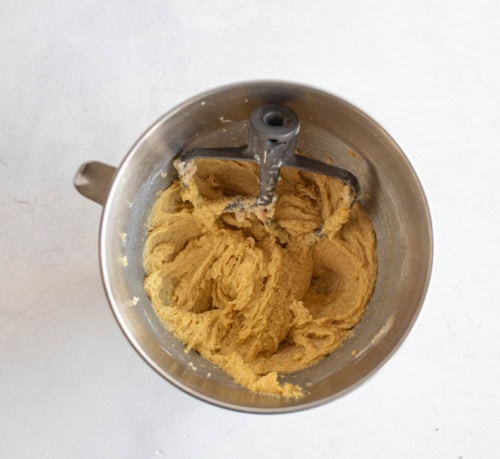 A metal mixing bowl with creamed cookie dough and a paddle attachment, viewed from above on a light-colored surface.