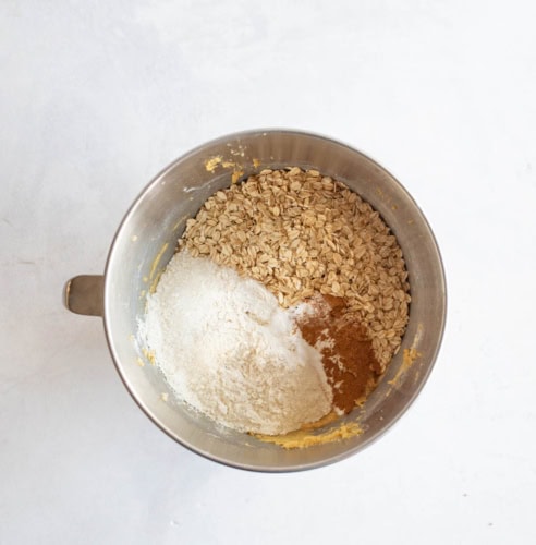 A metal mixing bowl containing rolled oats, flour, baking powder, and cinnamon, arranged in separate piles on a light surface.