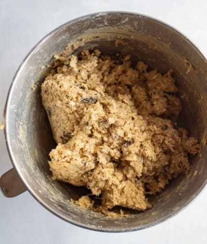 A metal mixing bowl filled with thick, chunky oatmeal cookie dough, featuring visible oats and chocolate chips.