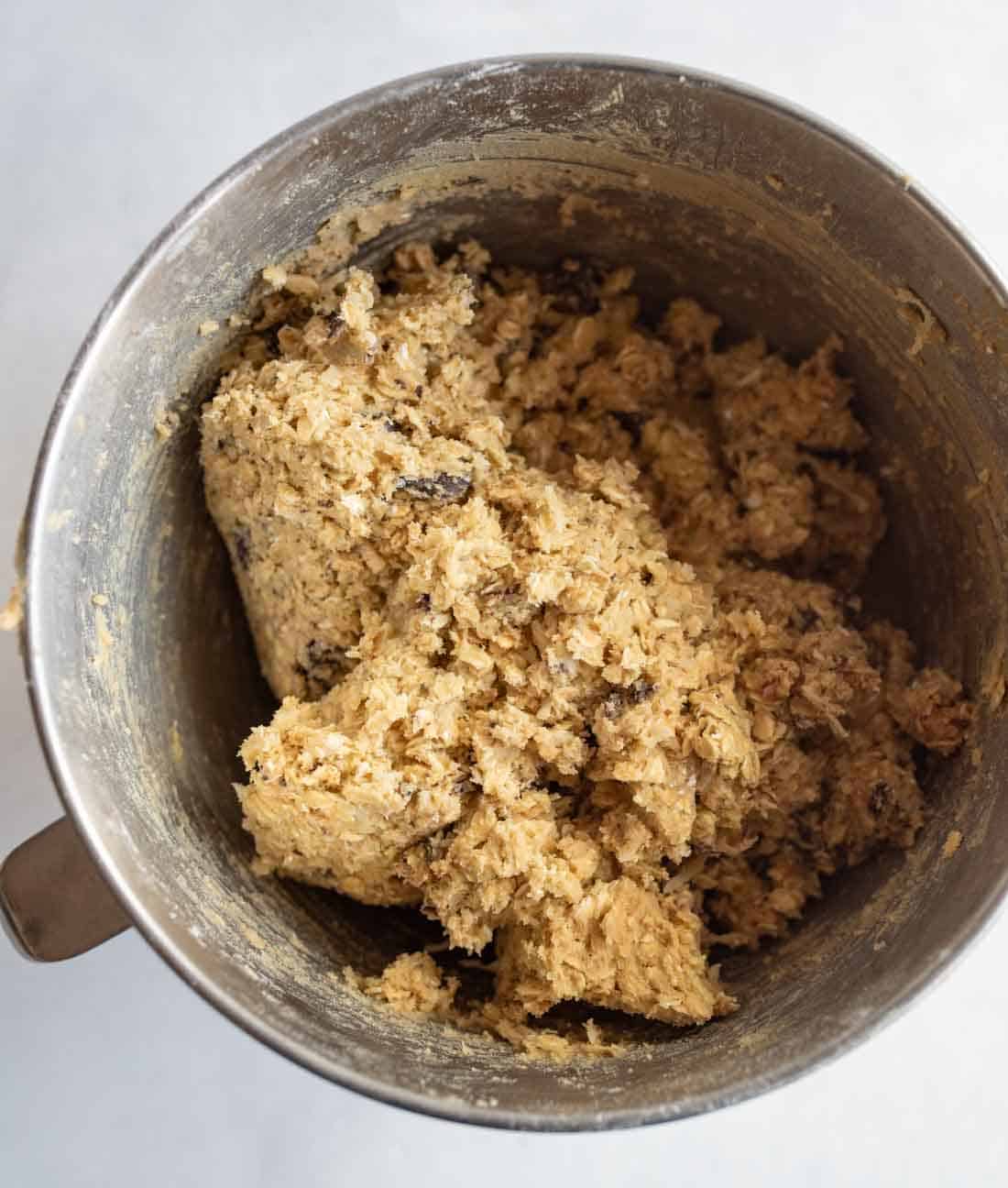 A metal mixing bowl filled with thick, chunky oatmeal cookie dough, featuring visible oats and chocolate chips.