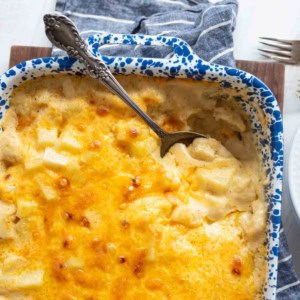 A blue-speckled baking dish filled with cheesy scalloped potatoes, topped with melted cheese. A silver serving spoon rests in the dish, and a blue-striped towel is nearby.