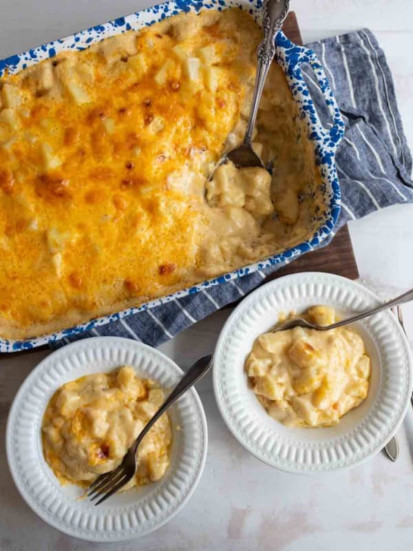 A blue and white speckled baking dish filled with cheesy potatoes, partially served into two white bowls with forks, all set on a striped cloth.