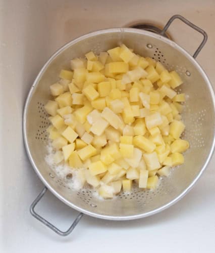 A metal colander filled with small, evenly diced raw potatoes sits in a white sink, with some water and foam visible around the potatoes.