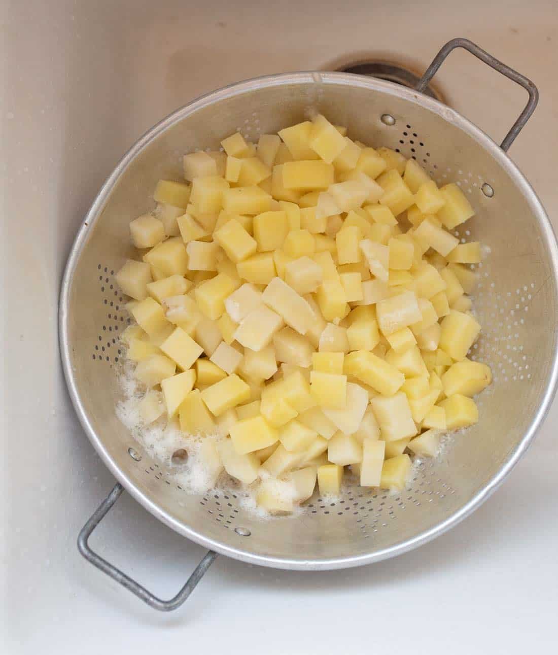 A metal colander filled with small, evenly diced raw potatoes sits in a white sink, with some water and foam visible around the potatoes.